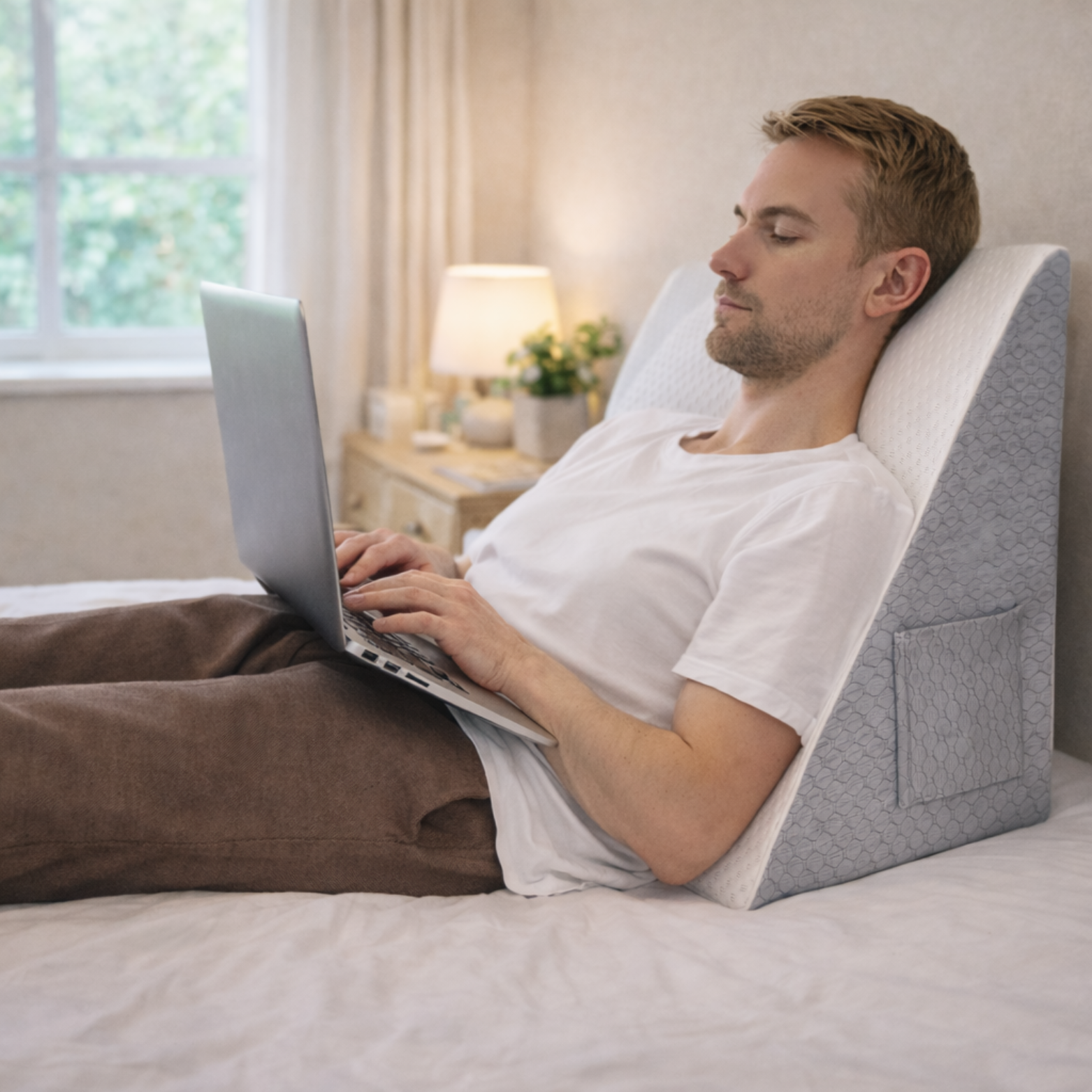 Homme utilisant un coussin ergonomique incliné pour soutenir le dos en travaillant sur un ordinateur portable dans un lit.